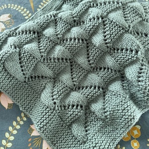 Op de afbeelding: Een zacht, saliegroen gebreid deken met een textuur, ruitvormig patroon.