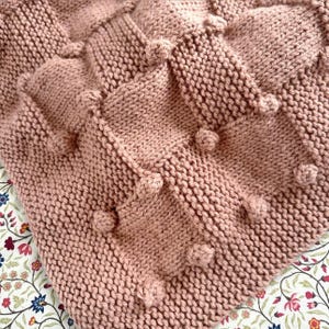 May include: A close-up of a knitted blanket in a soft rose colour. The blanket features a textured pattern with raised diamond shapes and small, round bobbles. The blanket is made of a thick yarn, perfect for warmth and comfort.