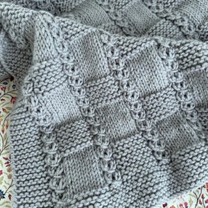 Puede incluir: Primer plano de una manta gris tejida con un patrón texturizado. La manta presenta un diseño repetitivo de cuadrados y puntadas decorativas. La manta suave y acogedora está hecha de hilo grueso y está doblada, mostrando sus intrincados detalles.