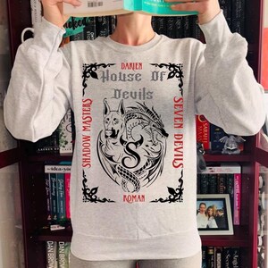 May include: A light gray sweatshirt with a graphic design featuring a dog, a dragon, and the text "DARIEN House Of Devils" in a gothic font. The words "SHADOW MASTERS" and "SEVEN DEVILS" are on the sides, and "ROMAN" is at the bottom.