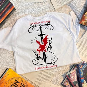 May include: White t-shirt with a black and red graphic design. The graphic features a sword with a dragon and the text "world ender ig morruthen".