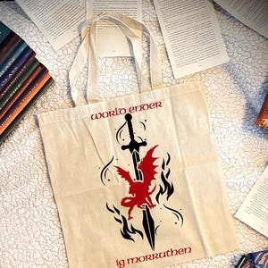 May include: A beige tote bag with the words "world ender" and "ig morruthen" in red. A black sword and red dragon graphic is centered on the bag. The bag has two white handles and is surrounded by open books.