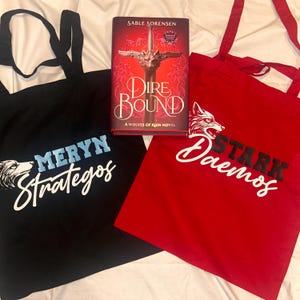 Op de afbeelding: Een boek getiteld "Dire Bound" van Sable Sorensen ligt tussen een zwarte draagtas met de tekst "Meryn Strategos" en een rode draagtas met de tekst "Stark Daemos". De tassen hebben wolf illustraties.