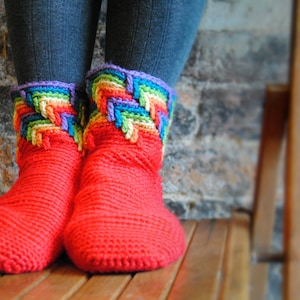 Puede incluir: Un par de botas de pantuflas de punto rojo brillante. La parte superior de las botas presenta un colorido diseño de arcoíris con ribete morado. Las botas están diseñadas para mantener los pies calientes.