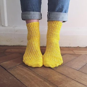 May include: A pair of bright yellow, open-weave socks are visible below rolled-up blue jeans. The socks have a textured, knitted appearance and are set against a backdrop of a wooden floor and a white wall.