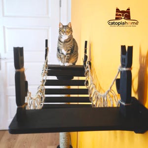 Può includere: Un albero per gatti nero con un ponte e una piattaforma. Il ponte è fatto di corda e la piattaforma ha un gatto nero seduto su di essa. L'albero per gatti è montato su un muro e ha un muro giallo dietro di esso. Il testo "catopiahome" è visibile nell'angolo in alto a destra dell'immagine.