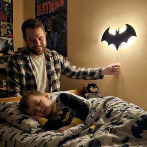Puede incluir: Una lámpara de pared negra con forma de Batman ilumina el dormitorio de un niño. La luz la sostiene un adulto, con un niño dormido en una cama con temática de Batman. La habitación presenta carteles y juguetes de Batman.