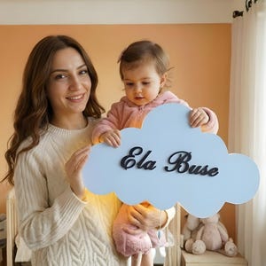 Könnte beinhalten: Eine hellblaue, wolkenförmige Wanddekoration mit dem Namen "Ela Buse" in schwarzer Schrift. Die Dekoration wird von einer Person und einem Kind gehalten. Der Hintergrund ist in einem warmen Pfirsichton gehalten.
