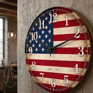 Op de afbeelding: Een ronde wandklok met een verweerd Amerikaans vlagontwerp in rood, wit en blauw. De wijzerplaat toont grote witte Arabische cijfers van 1 tot 12, zwarte uur- en minuutwijzers en de tekst EST. 1776 in het midden van de klok gedrukt.