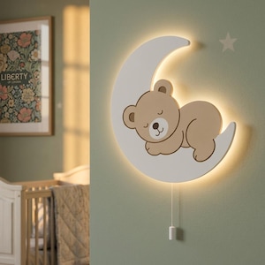 Könnte beinhalten: Eine LED-Wandleuchte in Form einer weißen Mondsichel mit einem schlafenden hellbraunen Bären darauf. Das Licht strahlt sanft hinter der Mondform hervor. Ein kleiner weißer Stern befindet sich über dem Mond und ein weißes Zugband hängt darunter.