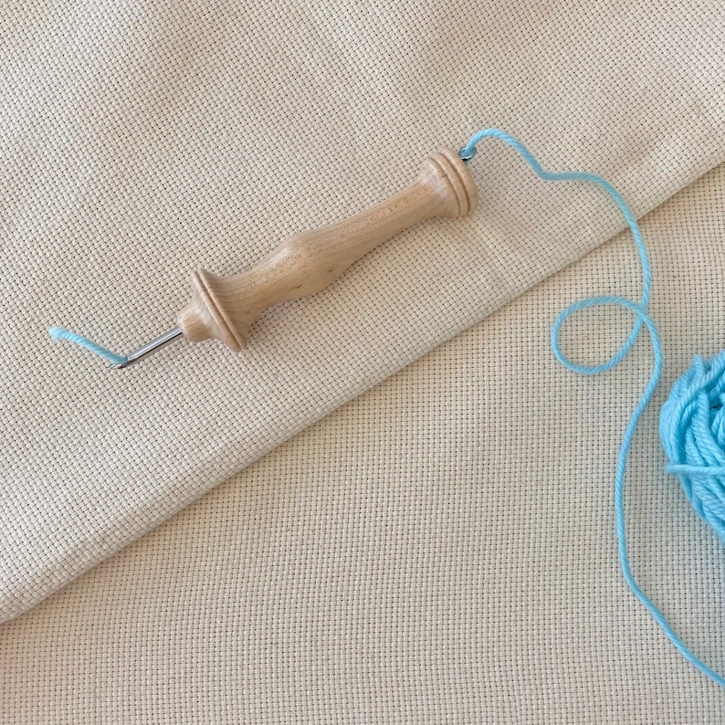 Puede incluir: Una herramienta de punto de cruz de madera con un hilo azul claro unido a ella. La herramienta est&aacute; sobre una tela de punto de cruz beige. Una bola de hilo azul claro est&aacute; en el fondo.