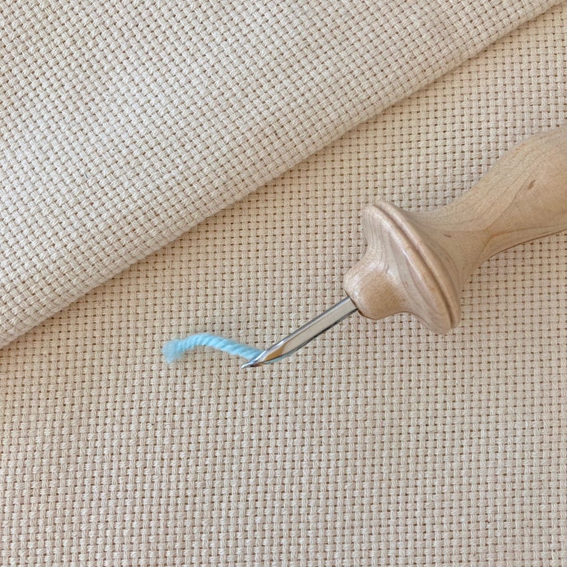 Puede incluir: Primer plano de un enhebrador de aguja de madera con un hilo azul claro en la aguja. El enhebrador est&aacute; sobre una tela Aida beige.