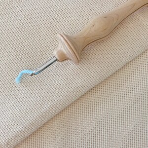 Puede incluir: Un enhebrador de aguja de punto de cruz de madera con un hilo azul claro. El enhebrador est&aacute; sobre un trozo de tela Aida beige.