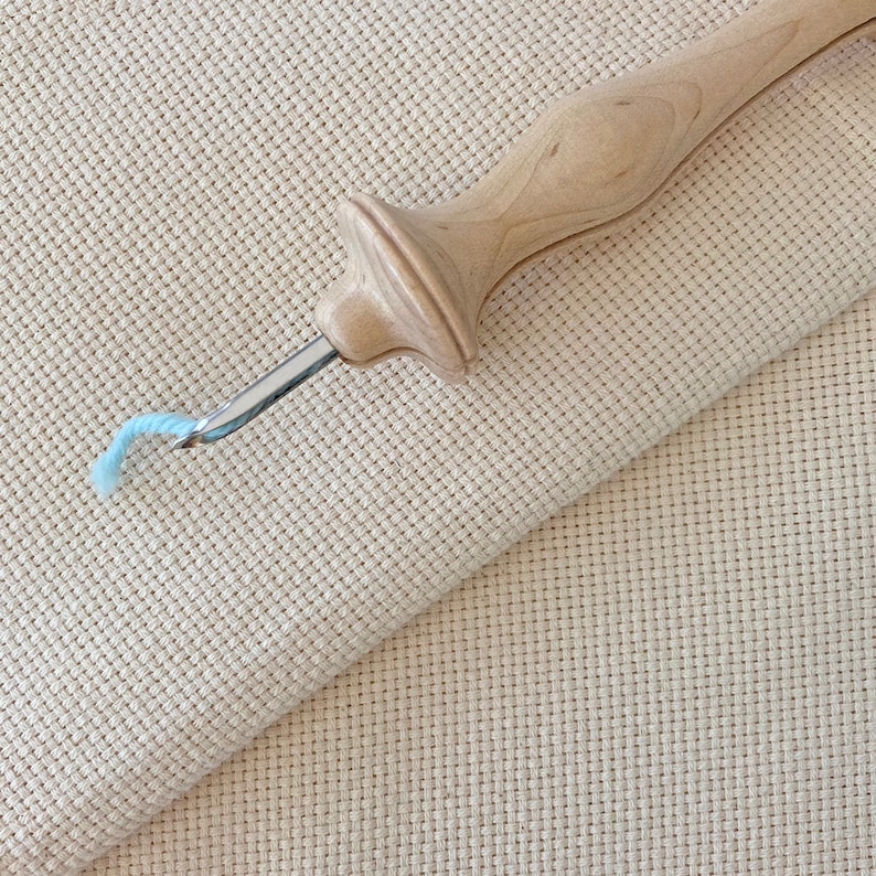 Puede incluir: Un enhebrador de aguja de punto de cruz de madera con un hilo azul claro. El enhebrador est&aacute; sobre un trozo de tela Aida beige.