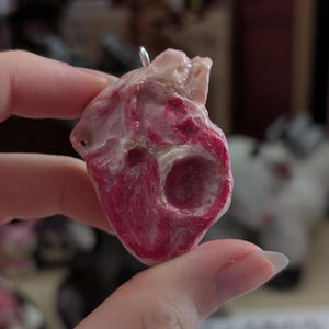 May include: A pendant shaped like an anatomical heart, featuring a textured surface with shades of pale pink, cream, and deep magenta. The piece has a glossy finish and includes a small silver-toned metal jump ring at the top for attachment.