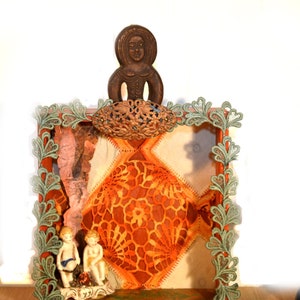 May include: A wooden frame with a green lace border, featuring a carved wooden figure, a patterned fabric background, and two porcelain figurines. The frame is decorated with a metal embellishment.