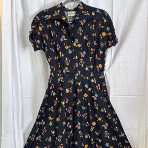 May include: A black vintage dress with a short sleeve and a full skirt. The dress is covered in a colorful pattern of small, whimsical illustrations. The dress has a button-up front and a collared neckline.