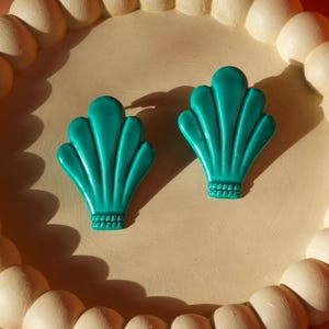 May include: Two teal seashell-shaped earrings on a cream-colored tray with a beaded edge. The earrings have a textured design and are set against a peach background. The tray is round and has a light beige color.