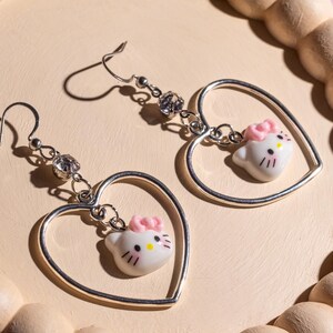 K&ouml;nnte beinhalten: Ein Paar silberner herzf&ouml;rmiger Ohrringe. Jeder Ohrring hat einen kleinen, wei&szlig;en Hello Kitty-Anh&auml;nger mit einer rosa Schleife und schwarzen Details, der im Herzen h&auml;ngt. Klare Kristallakzente sind ebenfalls vorhanden. Die Ohrringe werden auf einer hellrosa Oberfl&auml;che pr&auml;sentiert.