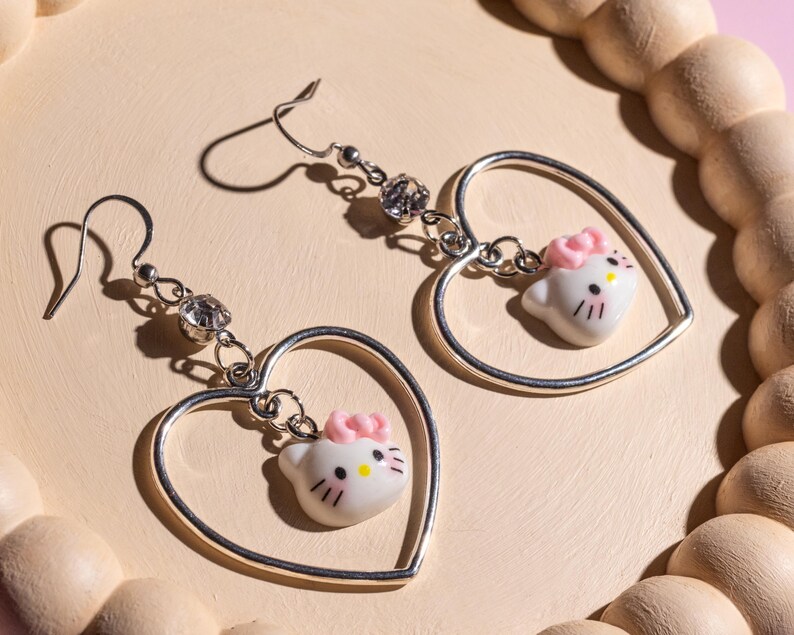 K&ouml;nnte beinhalten: Ein Paar silberner herzf&ouml;rmiger Ohrringe. Jeder Ohrring hat einen kleinen, wei&szlig;en Hello Kitty-Anh&auml;nger mit einer rosa Schleife und schwarzen Details, der im Herzen h&auml;ngt. Klare Kristallakzente sind ebenfalls vorhanden. Die Ohrringe werden auf einer hellrosa Oberfl&auml;che pr&auml;sentiert.