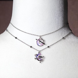 May include: A silver layered necklace with two charms. The charms are both crescent moons with a galaxy design. The top charm has a ring around it.