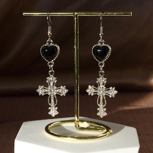 Könnte beinhalten: Ein Paar silberne Ohrhänger mit einem schwarzen Herz-Anhänger und einem Kreuz-Anhänger mit Strasssteinen.