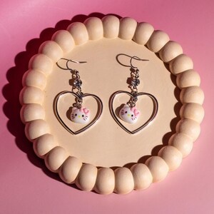 K&ouml;nnte beinhalten: Ein Paar silberfarbene H&auml;ngeohrringe. Jeder Ohrring hat einen herzf&ouml;rmigen Rahmen mit einem kleinen, wei&szlig;en Hello Kitty-Anh&auml;nger mit einer rosa Schleife. Die Ohrringe werden auf einem hellbeige, perlenbesetzten Tablett vor einem rosa Hintergrund pr&auml;sentiert.