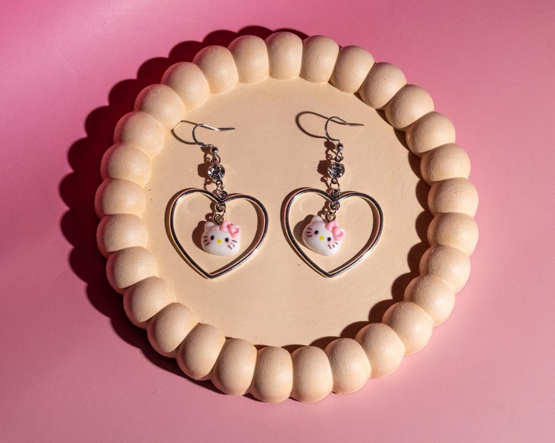 K&ouml;nnte beinhalten: Ein Paar silberfarbene H&auml;ngeohrringe. Jeder Ohrring hat einen herzf&ouml;rmigen Rahmen mit einem kleinen, wei&szlig;en Hello Kitty-Anh&auml;nger mit einer rosa Schleife. Die Ohrringe werden auf einem hellbeige, perlenbesetzten Tablett vor einem rosa Hintergrund pr&auml;sentiert.