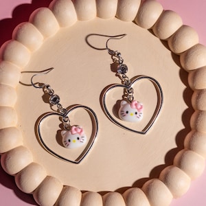 K&ouml;nnte beinhalten: Silberne herzf&ouml;rmige Ohrh&auml;nger mit einem wei&szlig;en Hello Kitty-Anh&auml;nger mit rosa Schleife. Die Ohrringe haben einen Strassstein-Akzent und werden auf einem hellbeige, perlenbesetzten Tablett vor rosa Hintergrund pr&auml;sentiert.