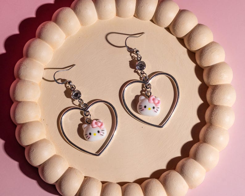 K&ouml;nnte beinhalten: Silberne herzf&ouml;rmige Ohrh&auml;nger mit einem wei&szlig;en Hello Kitty-Anh&auml;nger mit rosa Schleife. Die Ohrringe haben einen Strassstein-Akzent und werden auf einem hellbeige, perlenbesetzten Tablett vor rosa Hintergrund pr&auml;sentiert.