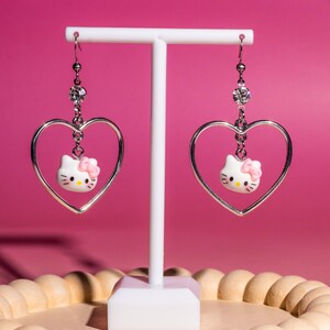 K&ouml;nnte beinhalten: H&auml;ngeohrringe mit einem silbernen herzf&ouml;rmigen Rahmen, einem wei&szlig;en Hello Kitty-Charakter mit rosa Schleife und einem Kristallakzent. Die Ohrringe h&auml;ngen an silbernen Haken. Sie werden auf einem wei&szlig;en St&auml;nder vor einem rosa Hintergrund pr&auml;sentiert.