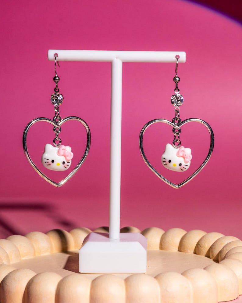 K&ouml;nnte beinhalten: H&auml;ngeohrringe mit einem silbernen herzf&ouml;rmigen Rahmen, einem wei&szlig;en Hello Kitty-Charakter mit rosa Schleife und einem Kristallakzent. Die Ohrringe h&auml;ngen an silbernen Haken. Sie werden auf einem wei&szlig;en St&auml;nder vor einem rosa Hintergrund pr&auml;sentiert.