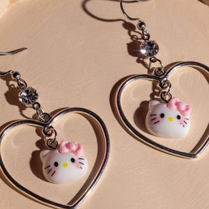 K&ouml;nnte beinhalten: Ein Paar silberfarbene Herz-Ohrh&auml;nger. Jeder Ohrring hat einen wei&szlig;en Hello Kitty-Anh&auml;nger mit einer rosa Schleife und einem funkelnden Strassstein-Akzent. Die Ohrringe sind an einem Fischhaken befestigt.