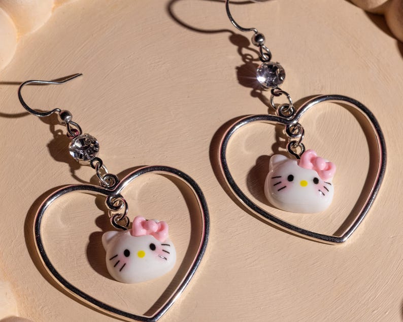 K&ouml;nnte beinhalten: Ein Paar silberfarbene Herz-Ohrh&auml;nger. Jeder Ohrring hat einen wei&szlig;en Hello Kitty-Anh&auml;nger mit einer rosa Schleife und einem funkelnden Strassstein-Akzent. Die Ohrringe sind an einem Fischhaken befestigt.