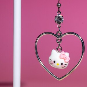 K&ouml;nnte beinhalten: Silberfarbene herzf&ouml;rmige Ohrringe mit einem wei&szlig;en Hello Kitty-Anh&auml;nger und einer rosa Schleife. Die Ohrringe h&auml;ngen an einem klaren Strassstein vor einem rosa Hintergrund. Ein s&uuml;&szlig;es Accessoire.
