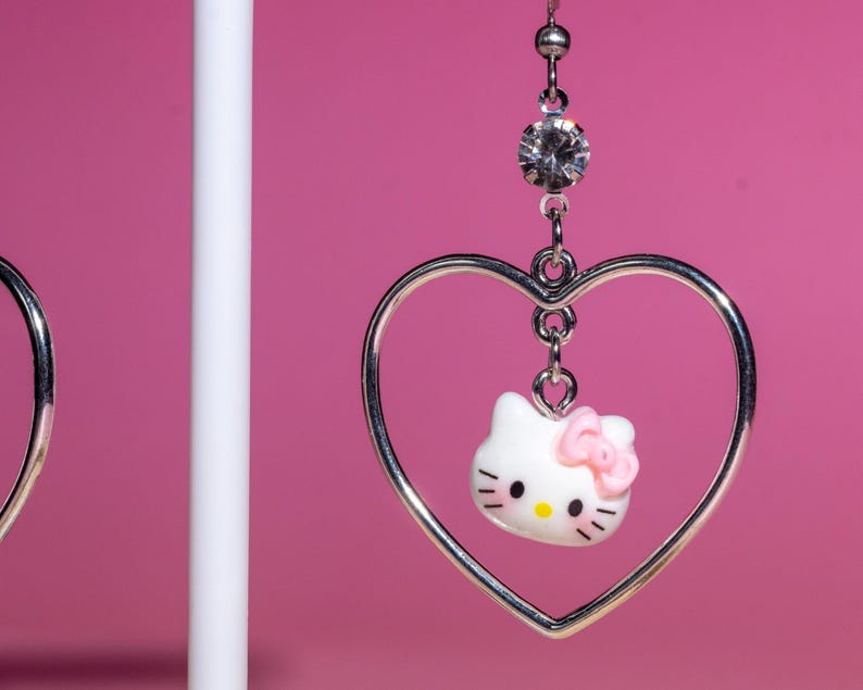 K&ouml;nnte beinhalten: Silberfarbene herzf&ouml;rmige Ohrringe mit einem wei&szlig;en Hello Kitty-Anh&auml;nger und einer rosa Schleife. Die Ohrringe h&auml;ngen an einem klaren Strassstein vor einem rosa Hintergrund. Ein s&uuml;&szlig;es Accessoire.