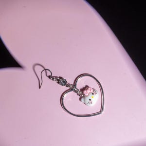 K&ouml;nnte beinhalten: Ein einzelner silberner Ohrring mit herzf&ouml;rmigem Rahmen und einem Hello Kitty-Anh&auml;nger. Der Anh&auml;nger hat eine rosa Schleife und ist an einer silbernen Kette mit einem kleinen Kristall befestigt. Der Ohrring befindet sich vor einem rosa herzf&ouml;rmigen Hintergrund.