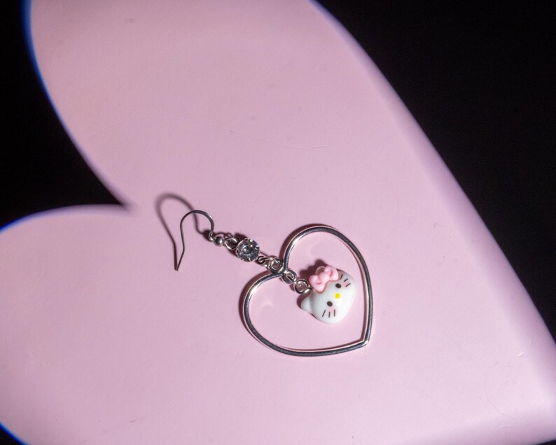 K&ouml;nnte beinhalten: Ein einzelner silberner Ohrring mit herzf&ouml;rmigem Rahmen und einem Hello Kitty-Anh&auml;nger. Der Anh&auml;nger hat eine rosa Schleife und ist an einer silbernen Kette mit einem kleinen Kristall befestigt. Der Ohrring befindet sich vor einem rosa herzf&ouml;rmigen Hintergrund.