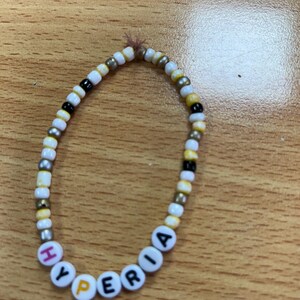 Puede incluir: Una pulsera de cuentas con la palabra "HYPERIA" escrita con cuentas de letras blancas. La pulsera presenta una mezcla de cuentas blancas, amarillas, negras y plateadas. La pulsera está sobre una superficie de madera.