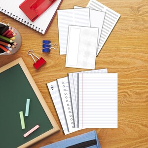 May include: An overhead shot of a wooden desk with various stationery items. There are several stacks of white paper with different line styles, a small chalkboard with chalk, and a pencil cup. A red binder clip and a blue binder clip are also visible.