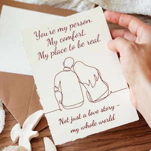 May include: A greeting card with a handwritten message and a line drawing of two people embracing. The text reads, "You're my person. My comfort. My place to be real. Not just a love story - my whole world."