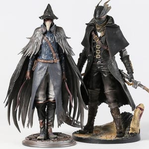 Puede incluir: Dos figuras detalladas de personajes de un videojuego. La figura de la izquierda lleva un sombrero negro, una larga capa gris y un abrigo azul. La figura de la derecha viste de negro, con un abrigo largo y una máscara. Ambas figuras están sobre bases detalladas.