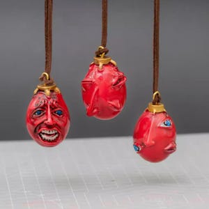 Könnte beinhalten: Drei rote, eiförmige Anhänger mit detaillierten Gesichtsmerkmalen und blauen Augen hängen an braunen Kordeln. Jeder Anhänger hat eine goldfarbene Kappe. Die Anhänger werden vor einem neutralen Hintergrund präsentiert.