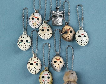 May include: A collection of ten miniature hockey mask keychains, each attached to a silver ball chain. The masks feature various finishes including white with black dots, metallic silver, and tan, with red triangular accents above the eyes.