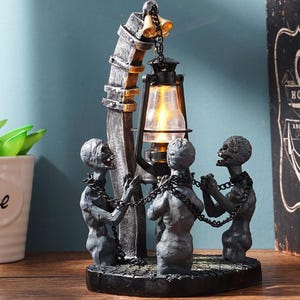 May include: A black metal lamp with a skeletal figure design. The lamp features a hanging lantern with a warm glow and three skeletal figures chained together. The lamp is on a wooden surface with a book that says "Hope Anchors the Soul".