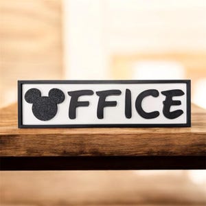 Könnte beinhalten: Ein schwarz gerahmtes Schild mit dem Wort "OFFICE" in schwarzen Buchstaben. Auf der linken Seite des Schildes befindet sich ein schwarzer, glitzernder Mickey Mouse-Kopf. Der Hintergrund des Schildes ist weiß. Das Schild steht auf einer Holzoberfläche.