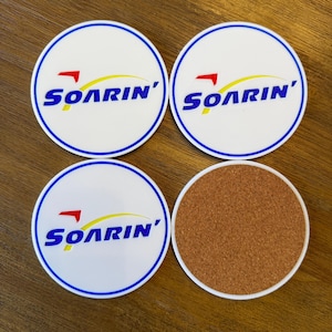 May include: Four round coasters with a white background and blue border. Three coasters feature the word "Soarin'" in blue and yellow letters with a red airplane icon. The fourth coaster is brown and cork.