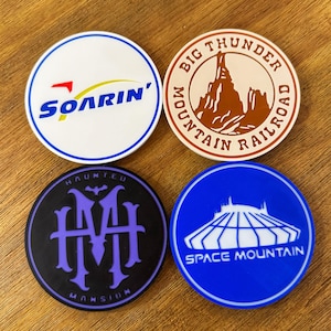 Peut inclure: Quatre dessous de verre ronds avec des motifs de parcs à thème. Ils présentent "Soarin'" en bleu et rouge sur blanc, "Big Thunder Mountain Railroad" en marron sur beige, "Haunted Mansion" en violet sur noir et "Space Mountain" en blanc sur bleu.