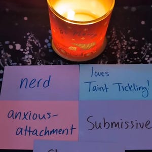 May include: Four pink, blue, and purple sticky notes with handwritten text. The notes say "nerd", "loves taint tickling", "anxious-attachment", and "submissive". A fifth sticky note says "strong pull-out game". A lit candle with a red and white design is in the background.
