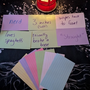 May include: A set of eight index cards with handwritten text. The cards are in various colors, including yellow, green, pink, purple, and blue. The text on the cards includes "nerd", "loves spaghetti", "3 inches (soft)", "recently broke a bone", "wipes back to front", and "'Straight'".